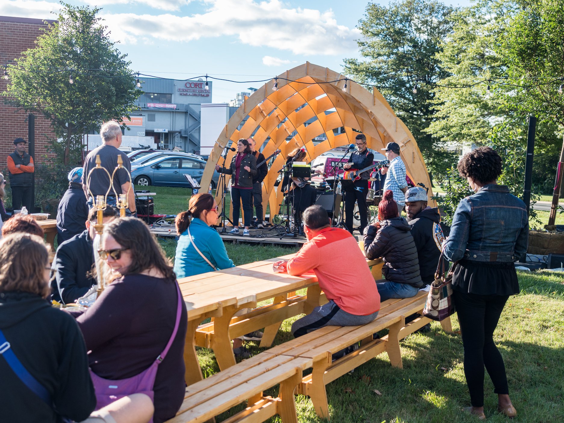 People sit on yellow picnic benches watching a small band perform on an outdoor stage under a wooden arch structure on a sunny day.