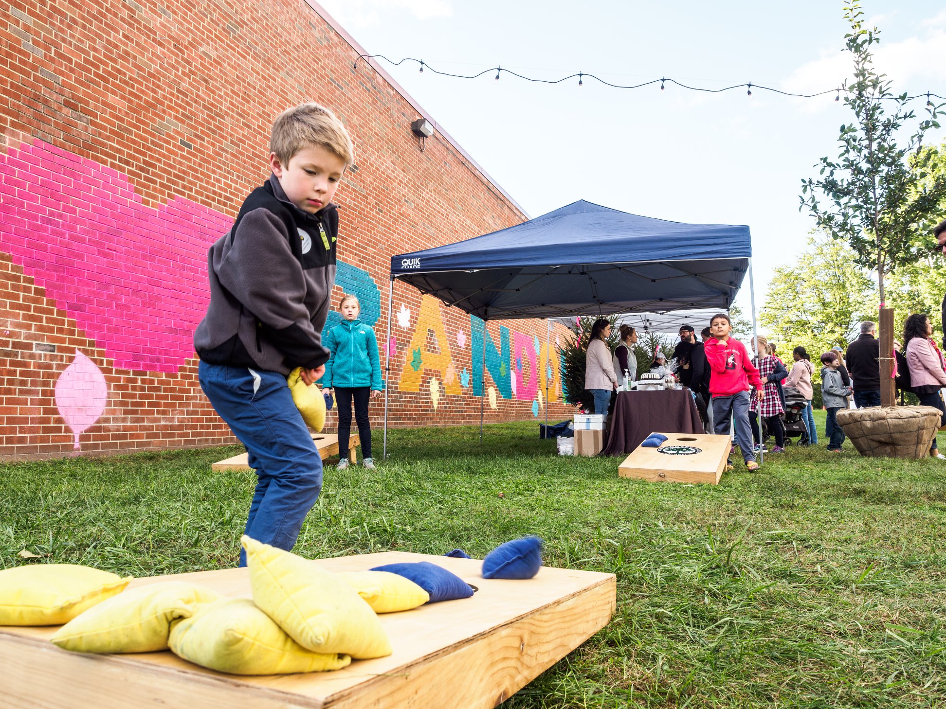 A young boy plays a bean bag toss game outdoors near a brick wall mural, with people gathered under a canopy in the background.