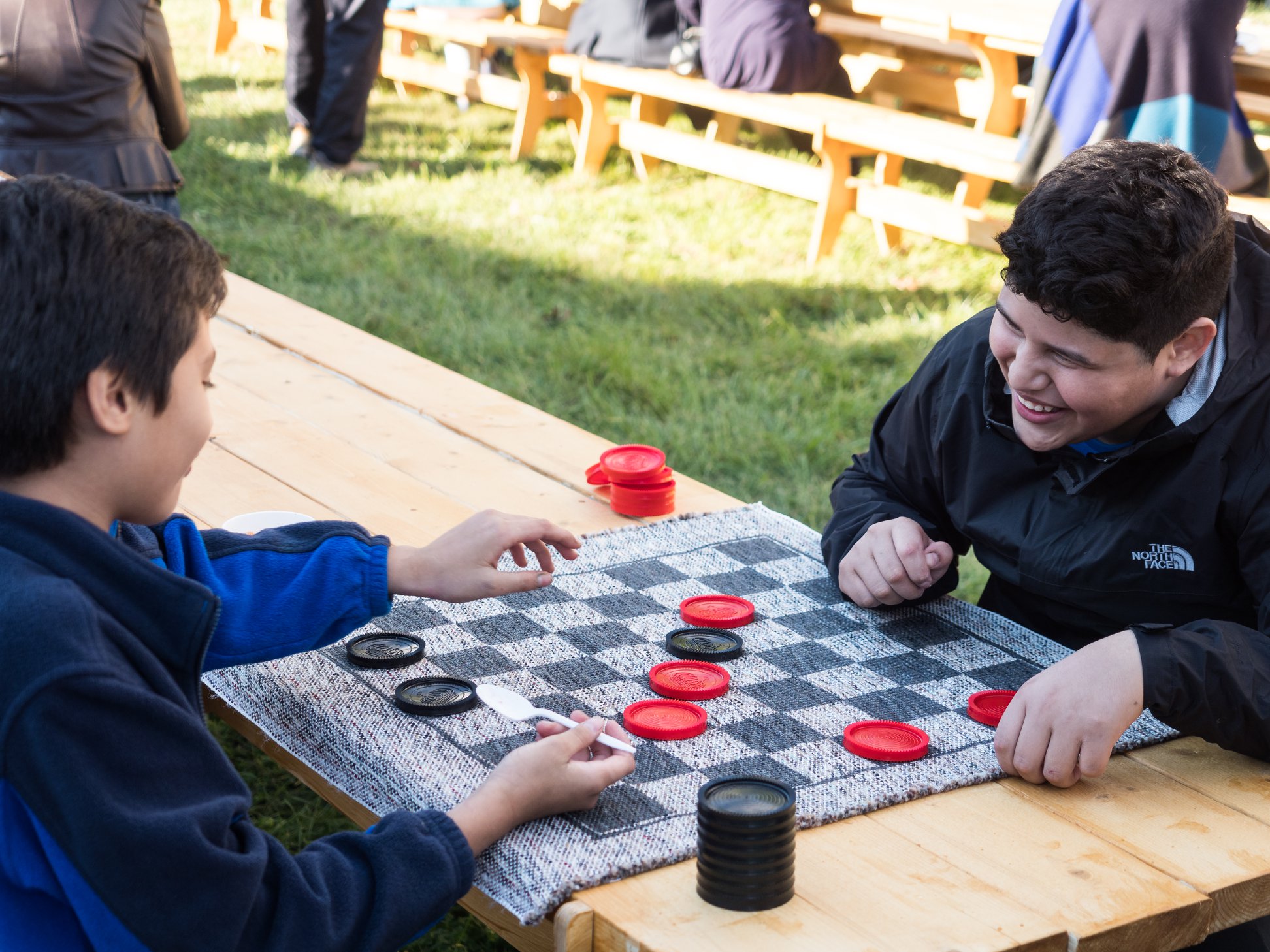 Two boys sit at a wooden picnic table outdoors, playing a game of checkers and smiling.