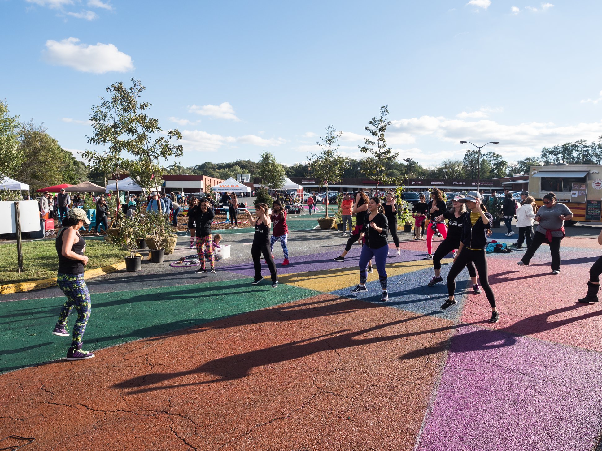 A group of people participate in an outdoor fitness class on a painted pavement in a park-like area under a clear sky.