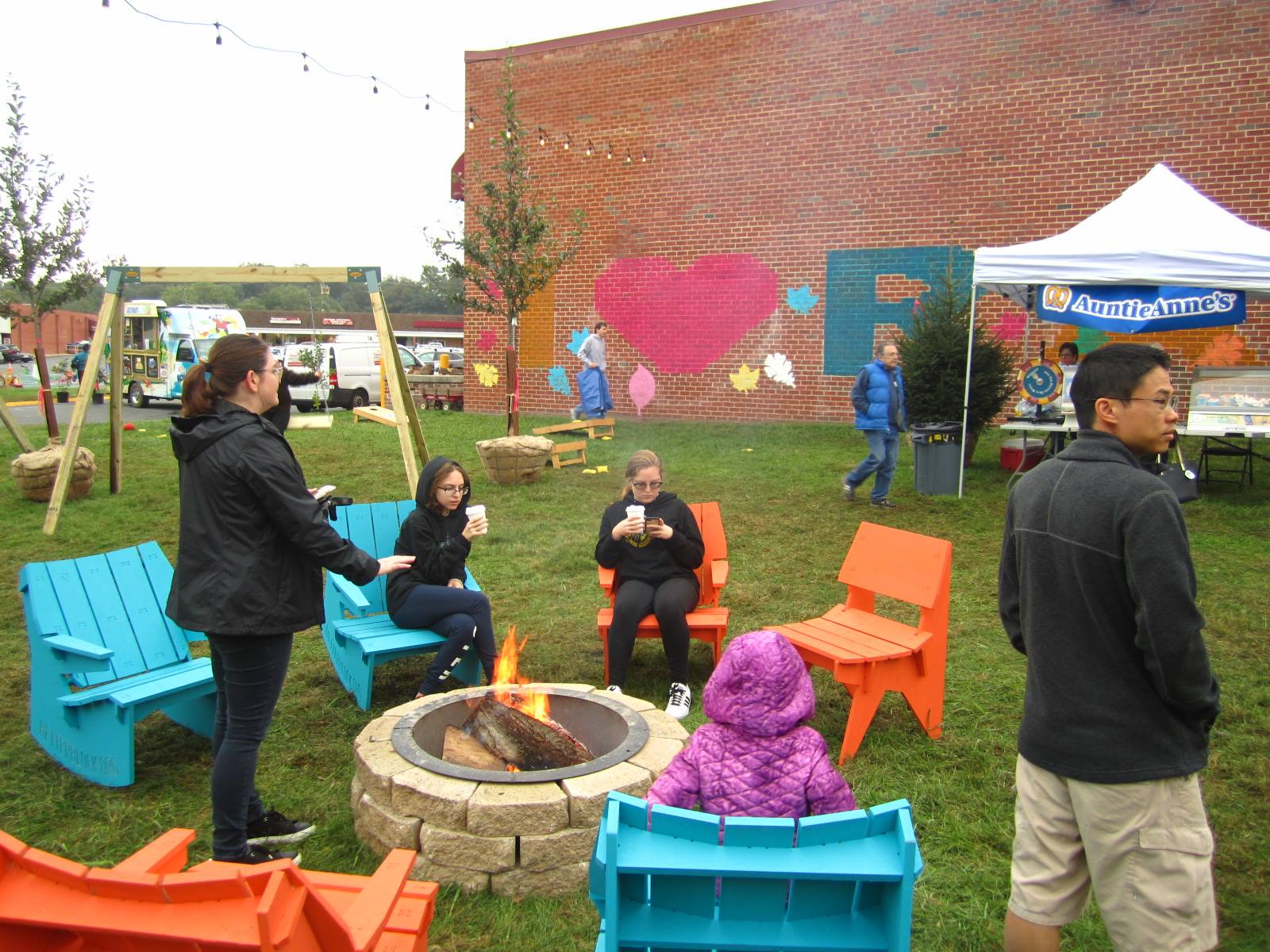 People sit on colorful chairs around a fire pit at an outdoor event; a mural with a large pink heart is on a brick wall in the background.