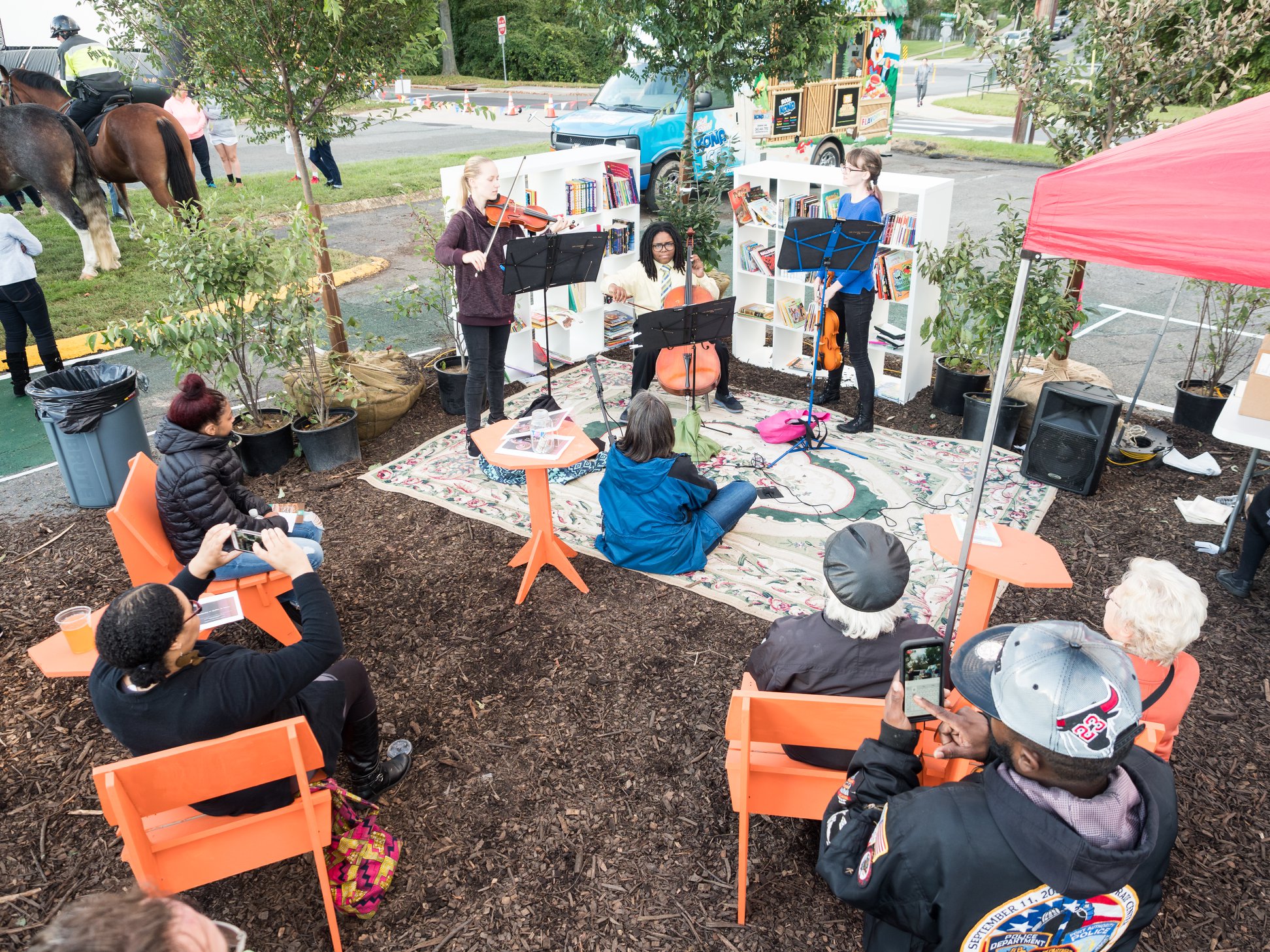 A small string quartet performs outdoors on a rug while people sit on orange chairs and take photos; trees, bookshelves, and a tent surround the area.