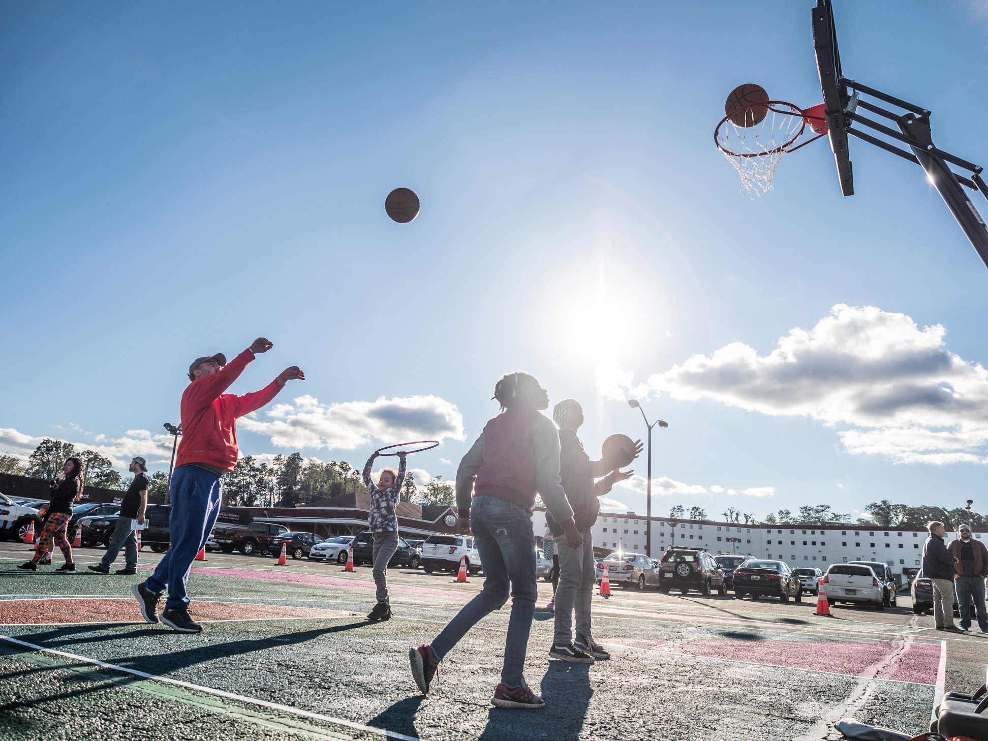 A group of people play basketball on an outdoor court under a sunny sky, with cars parked nearby and a building in the background.
