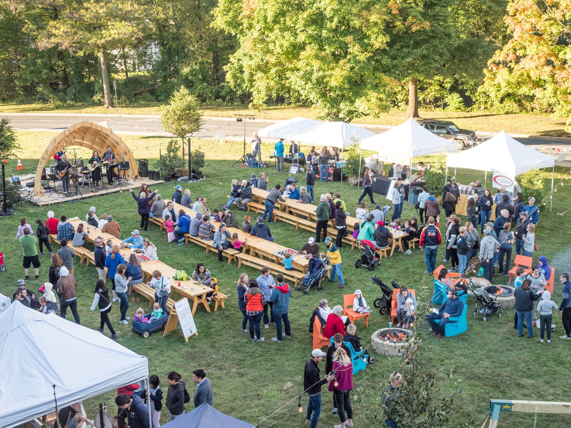 A community outdoor event with people gathered at picnic tables, tents, and a small stage, surrounded by trees on a sunny day.