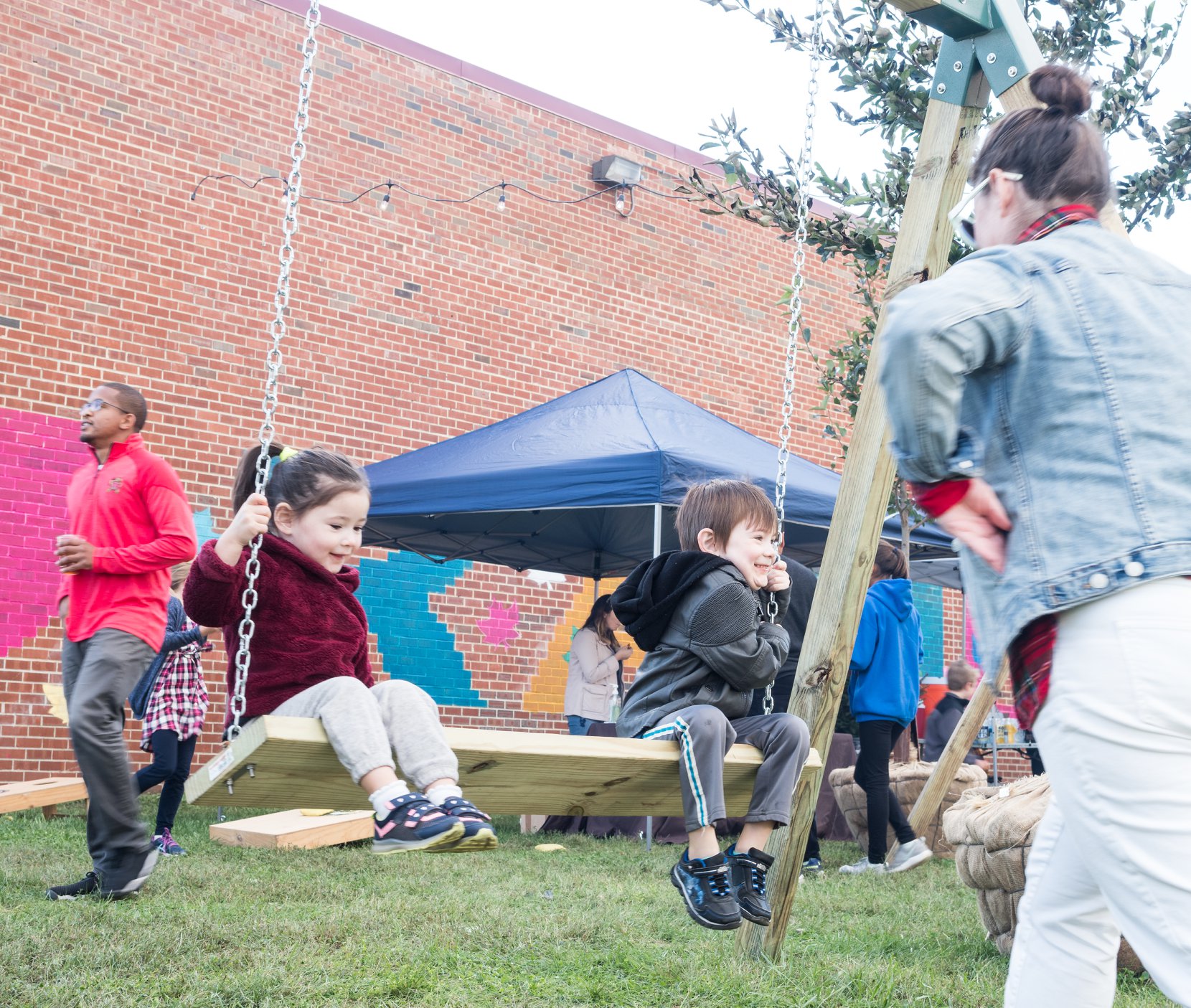 Two children sit on a wooden swing while an adult stands nearby; other people and a blue tent are visible in the background.
