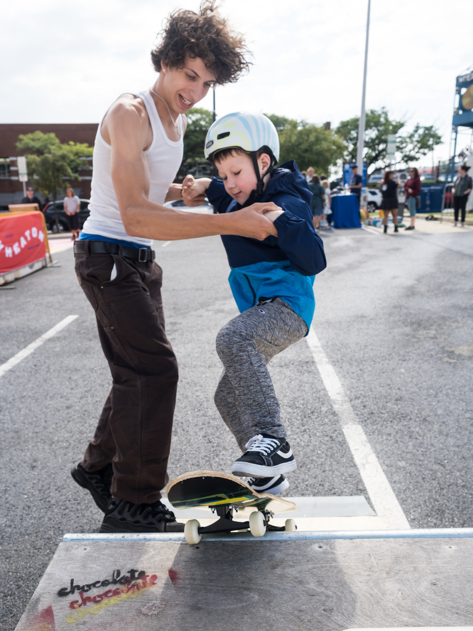 A young man helps a child wearing a helmet and blue jacket balance on a skateboard at an outdoor skate park event.