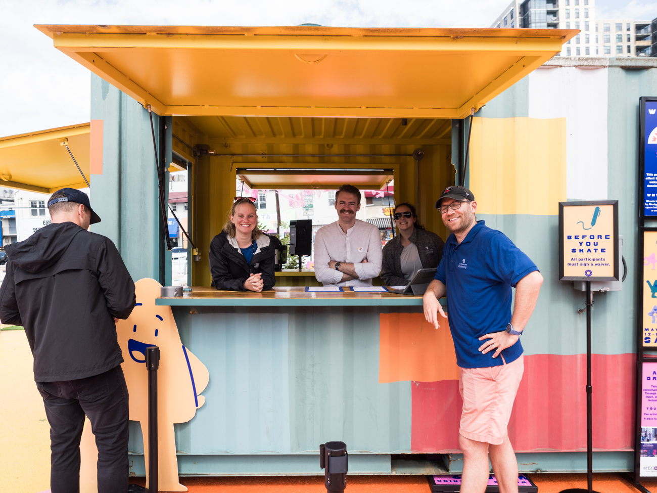 Five people are gathered at an outdoor service kiosk made from a shipping container, with two people standing outside and three people standing inside behind the counter.