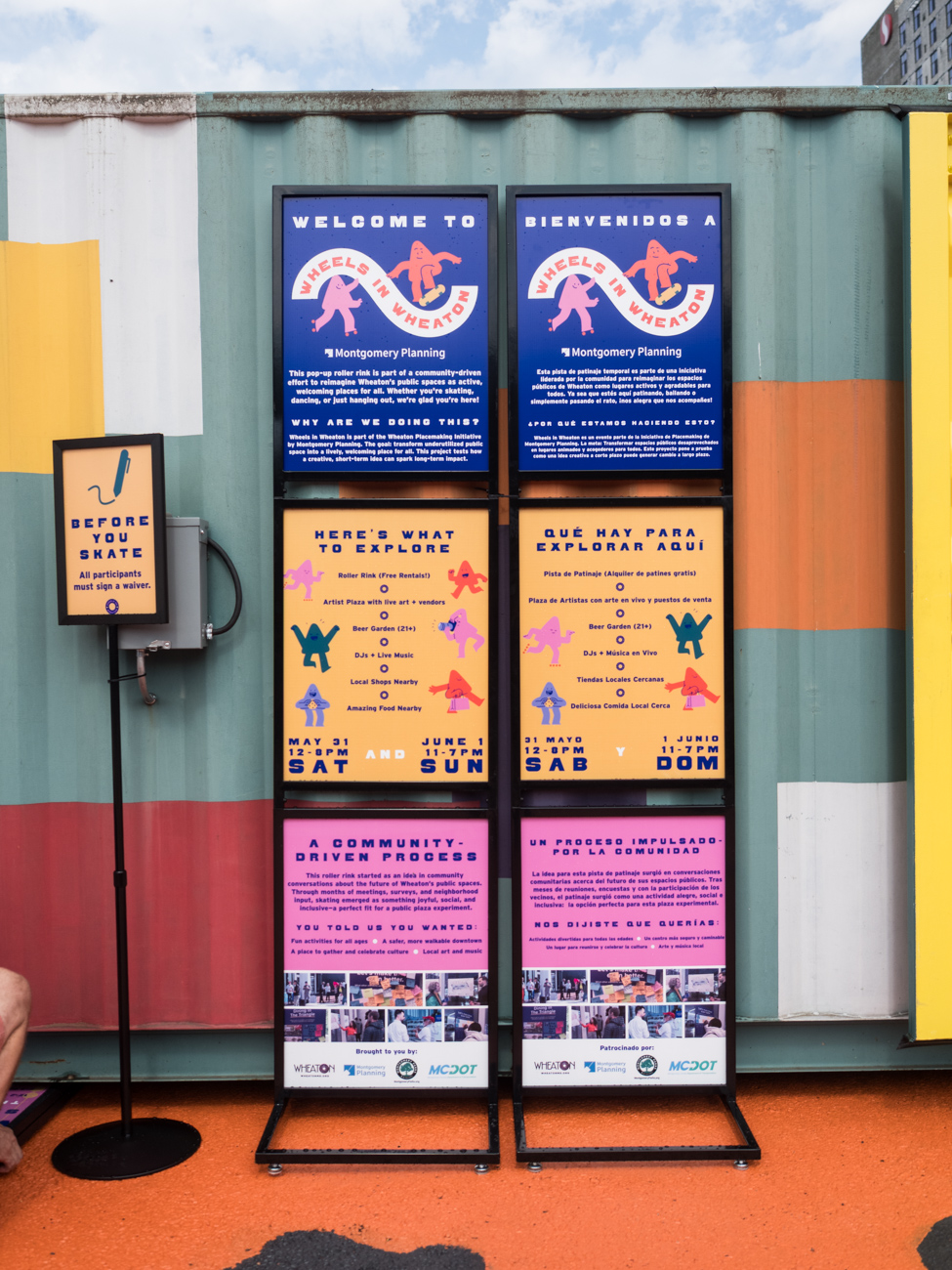 Information boards in English and Spanish display event details, activities, and community programs in front of a colorful wall. A smaller sign stands on the left.