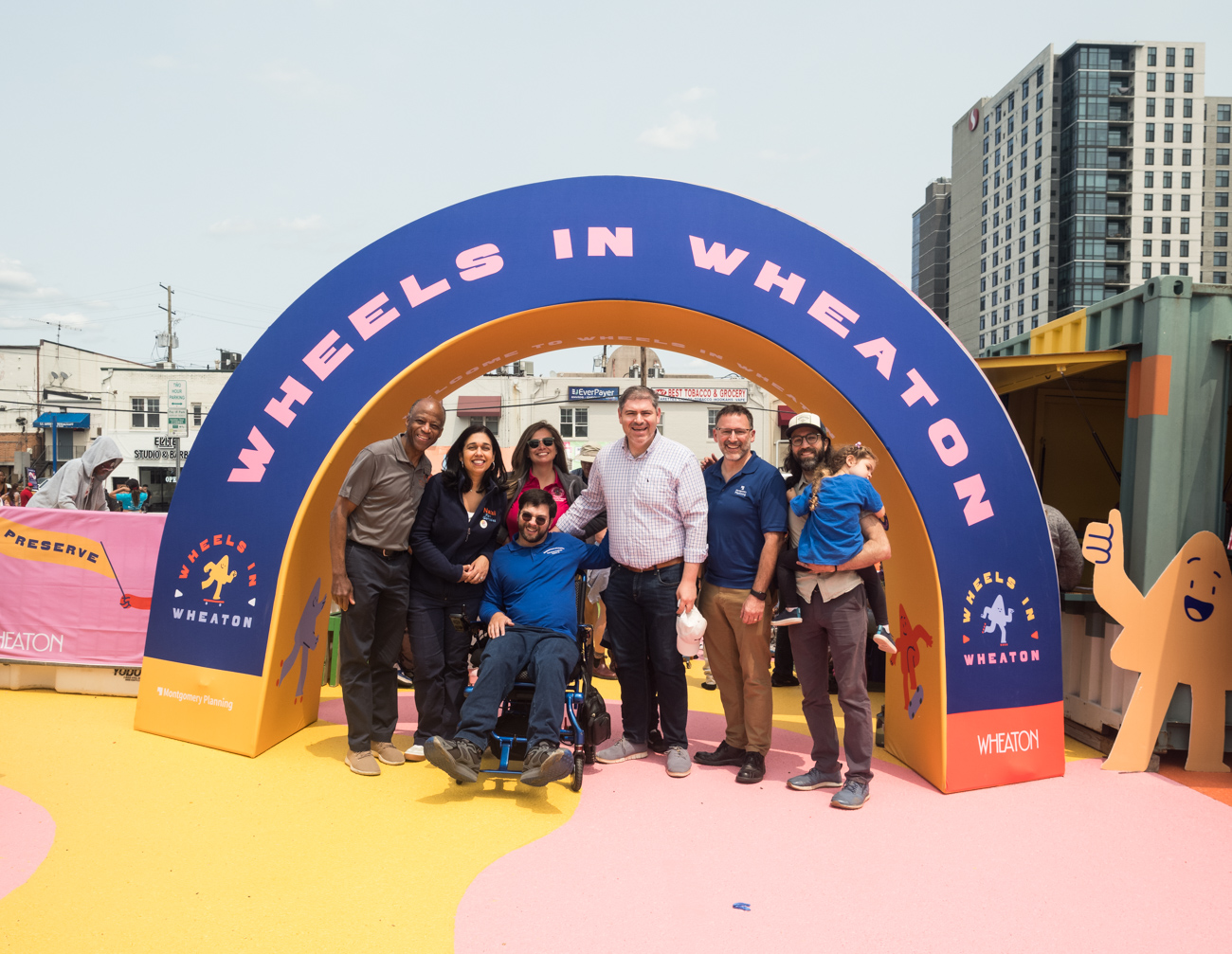 A group of seven adults and one child pose and smile under an archway labeled "Wheels in Wheaton" at an outdoor event in a city setting.