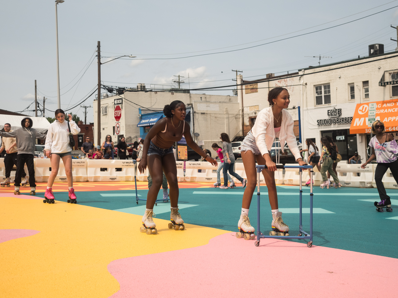 People roller skating on a colorful outdoor rink, with some using walkers for support; others and onlookers are visible in the background.