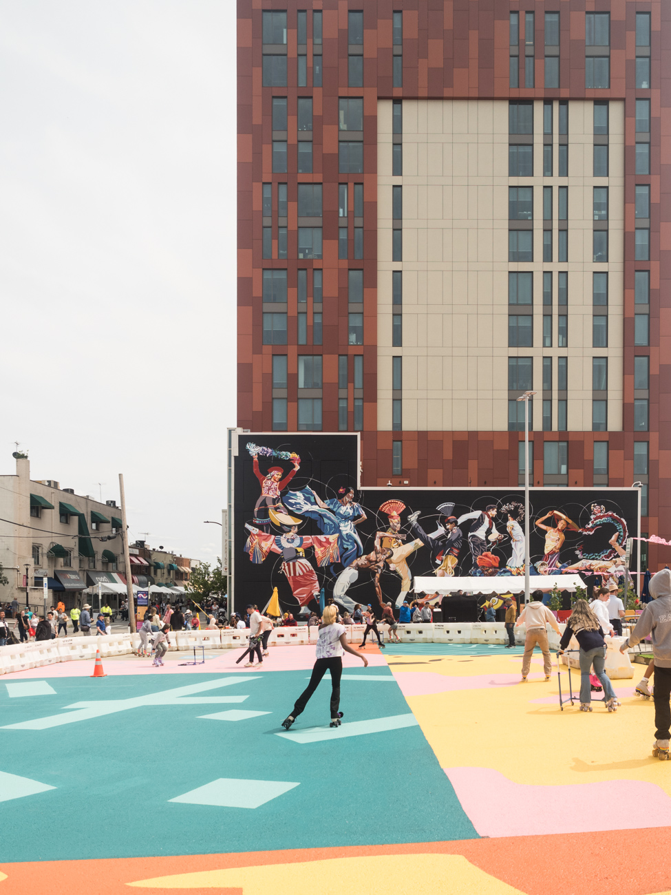 People roller skating on a colorful outdoor rink in an urban area, with a large mural and a tall building in the background.