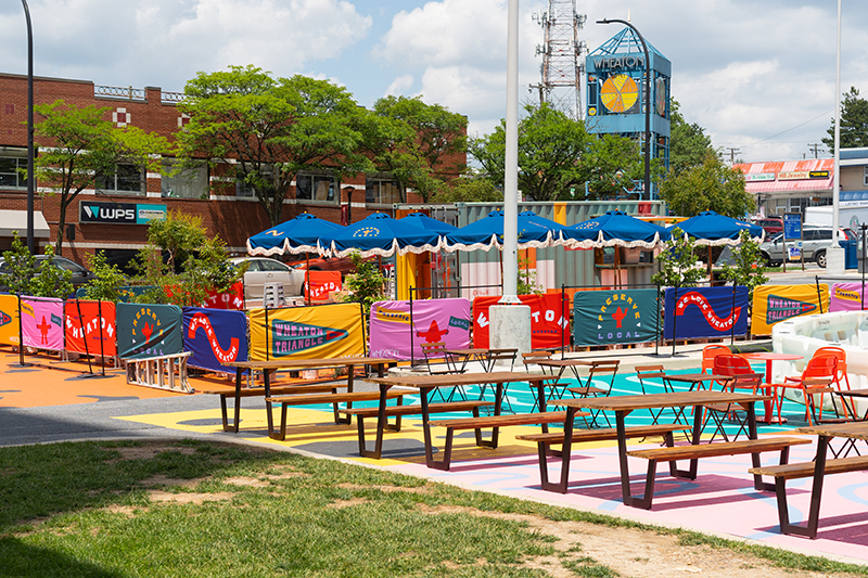 Outdoor seating area with wooden picnic tables and orange chairs, colorful painted ground and barriers, umbrellas, and nearby buildings in an urban setting.