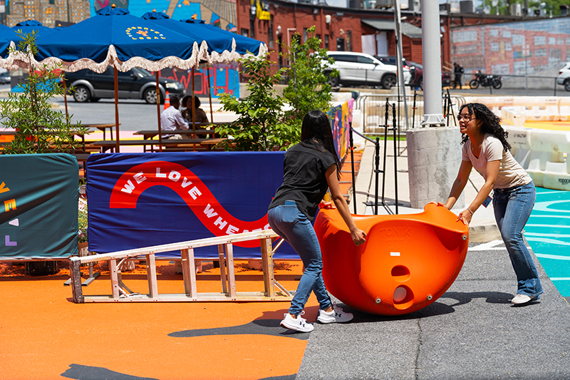Two women move a large orange play structure in an outdoor urban space with colorful seating, banners, and painted pavement.