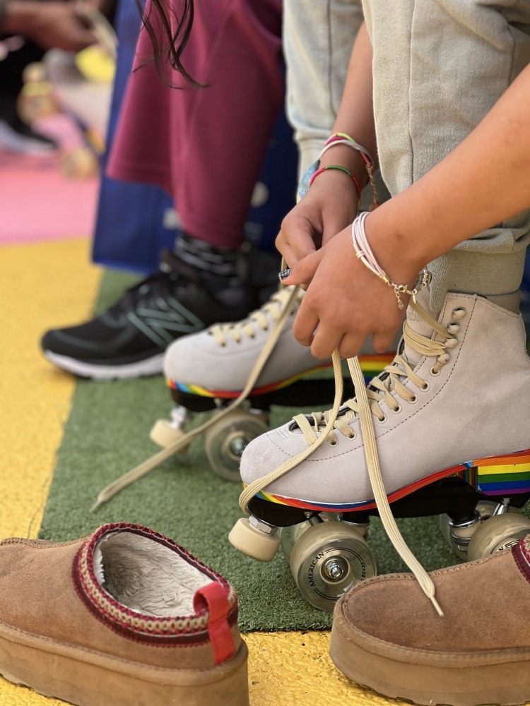 A person ties the laces on white roller skates with rainbow details; another person wearing a sneaker is nearby, and a slipper is on the ground.