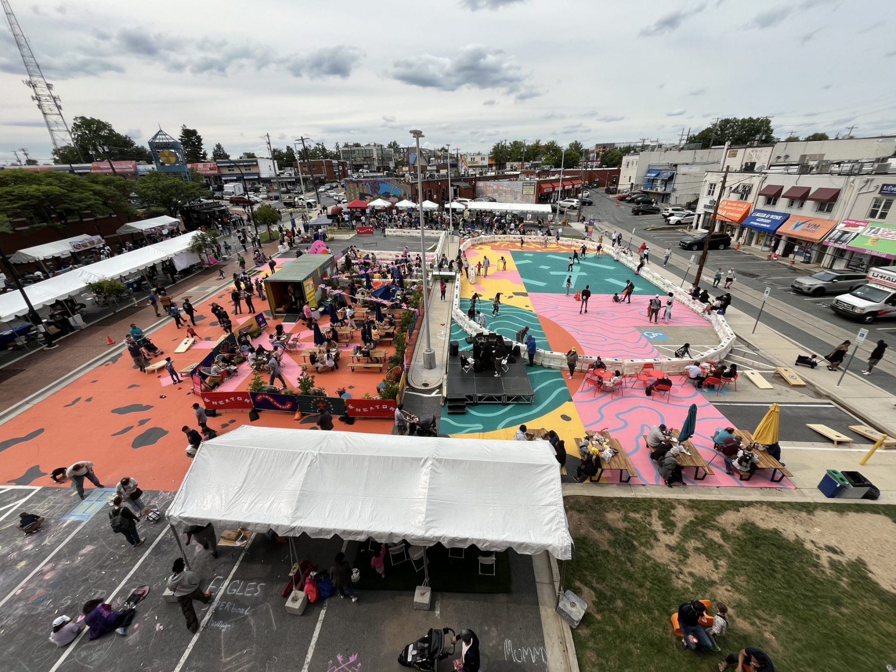 Aerial view of a public plaza with colorful painted ground, tents, people walking, and vendors set up along the edges on a cloudy day.