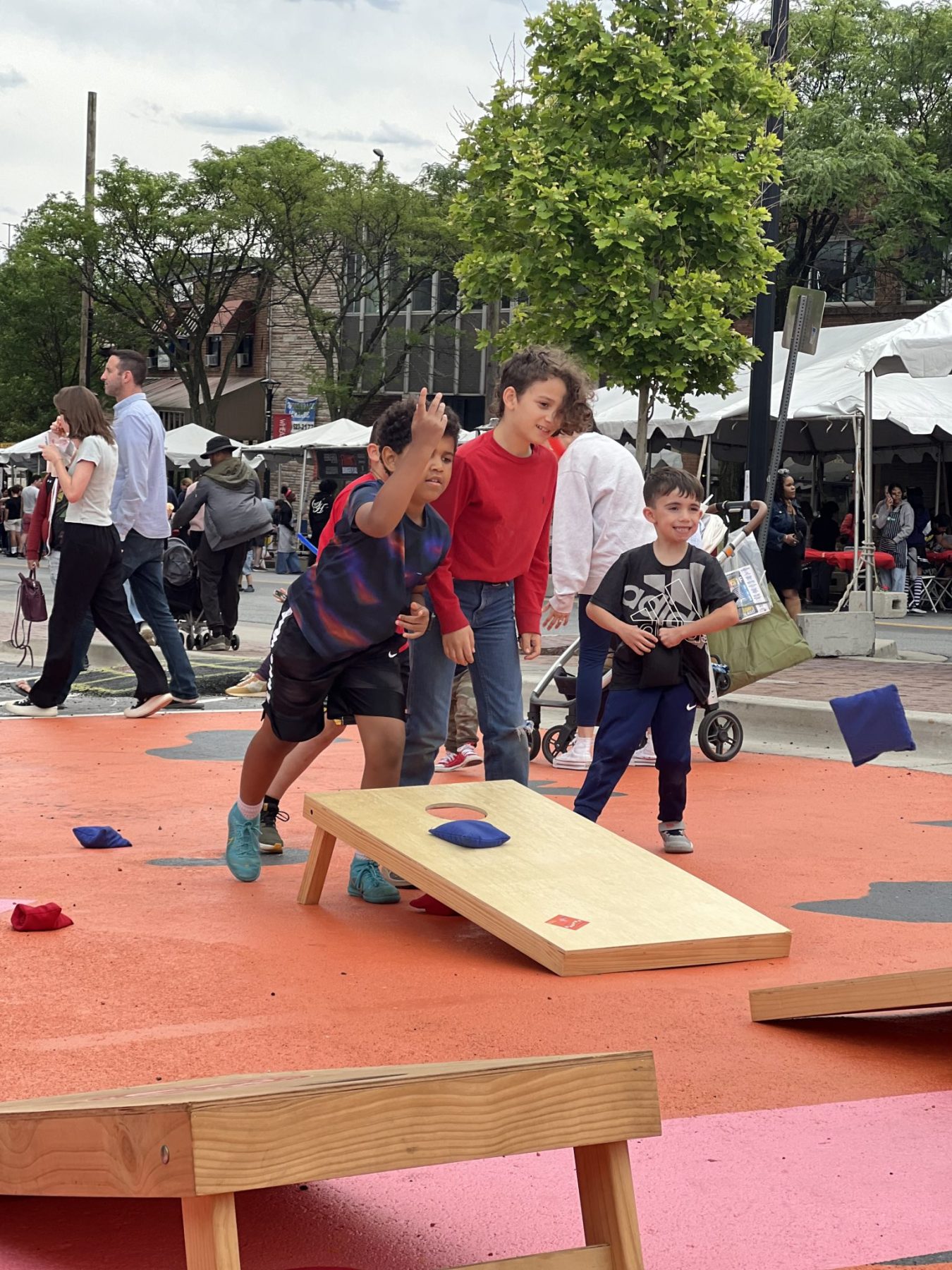 Children play a bean bag toss game on an outdoor painted surface while adults and other children watch in a busy public area.