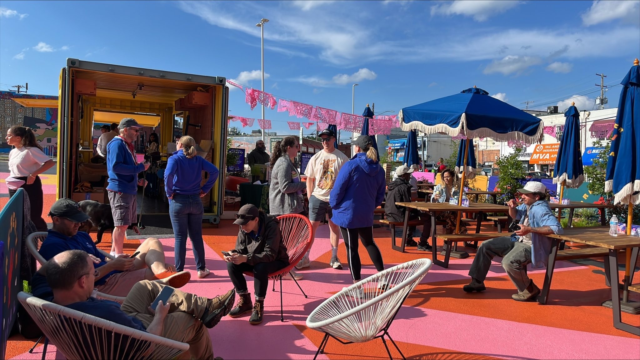 People sit and stand in an outdoor space with colorful flooring, umbrellas, and decorations under a blue sky. Some converse, others use phones or relax on chairs.