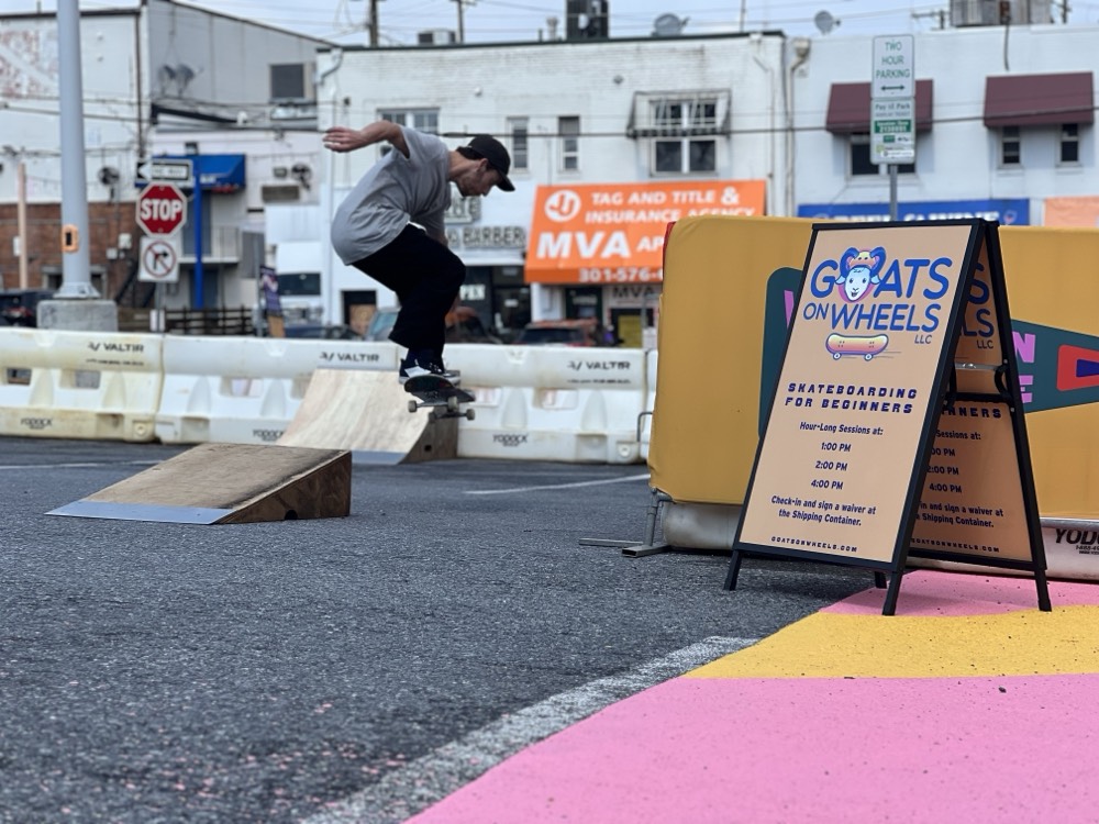 A skateboarder performs a trick off a wooden ramp in a parking lot near a “Goats on Wheels” skateboarding for beginners sign. Temporary barriers and businesses are in the background.