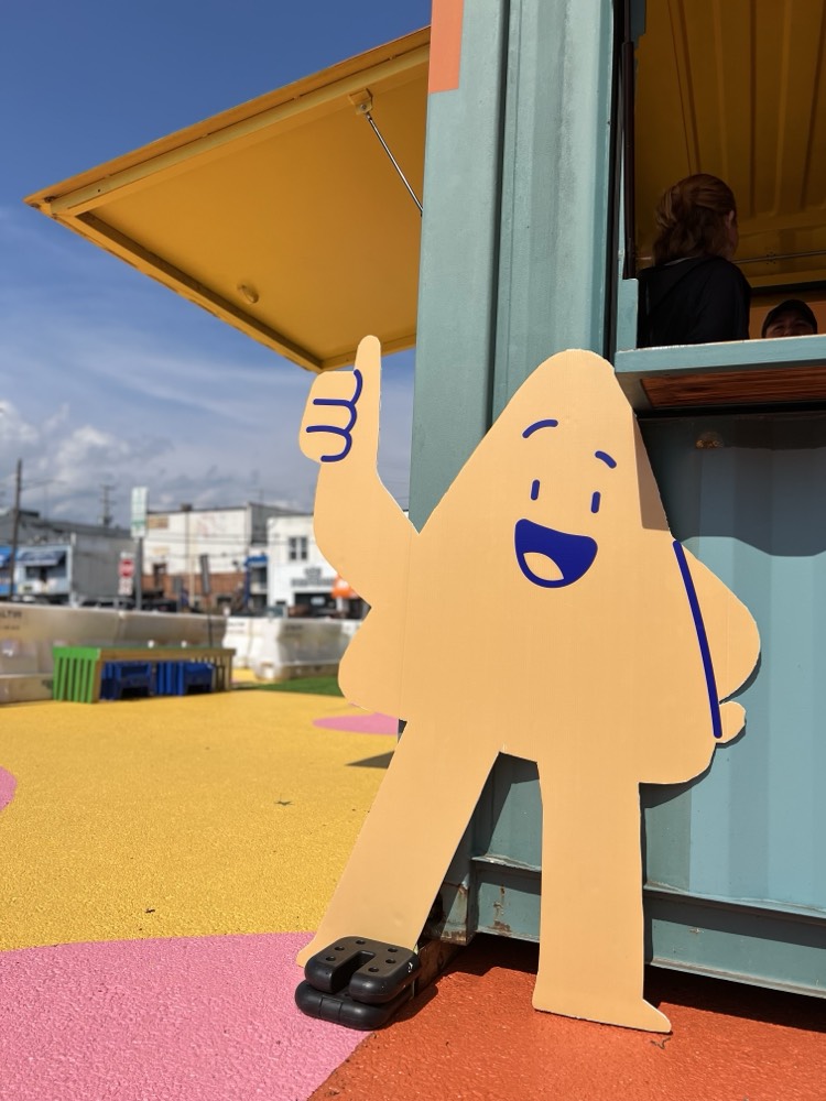 A large, yellow, cartoon-like cutout with a smiling face and a thumbs-up leans against a colorful outdoor kiosk on a sunny day.