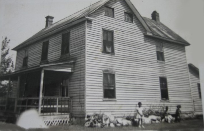 Black and white photo of a two-story wooden house with a porch; two children and several goats stand near the side of the building.