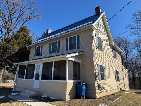 Two-story beige house with a blue metal roof, screened front porch, blue trash bin, and a small snow patch on the lawn under a clear blue sky.
