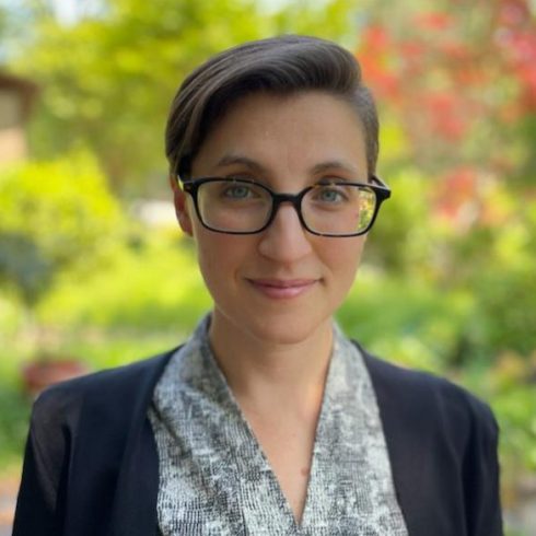 Person with short brown hair and glasses, wearing a patterned blouse and black blazer, standing outdoors with greenery and red flowers blurred in the background.