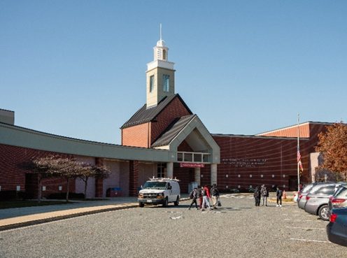 A brick school building with a central steeple, a parked van, and several people walking in the parking lot under a clear blue sky.