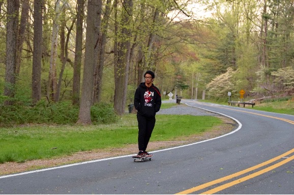 Sligo Creek Open Parkway
A person rides a skateboard on a winding road through a forested area, wearing dark clothing and glasses, with hands in pockets.