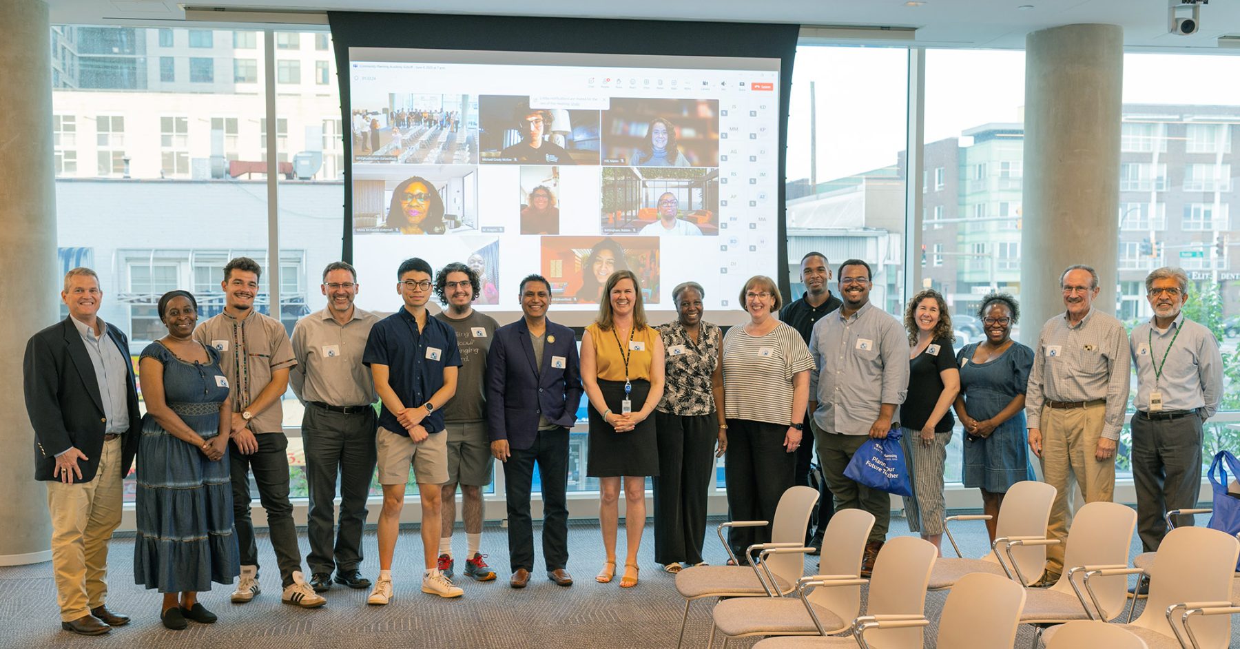 A group of people stands in a conference room in front of a screen displaying a virtual meeting with multiple participants.