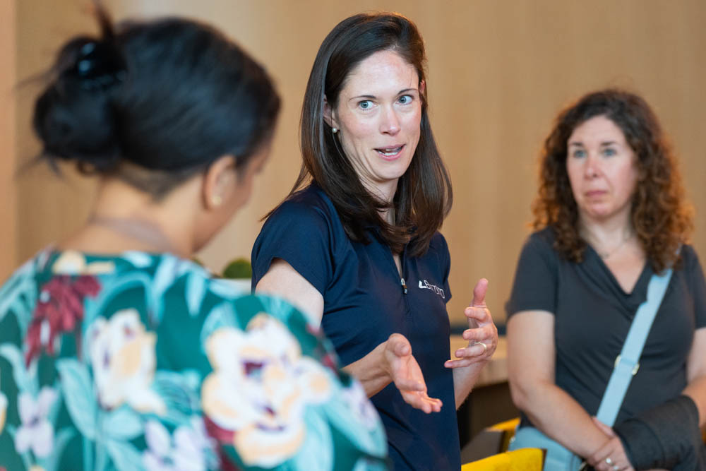 Three women are having a conversation indoors; one woman in the center is gesturing with her hands while speaking to the others.