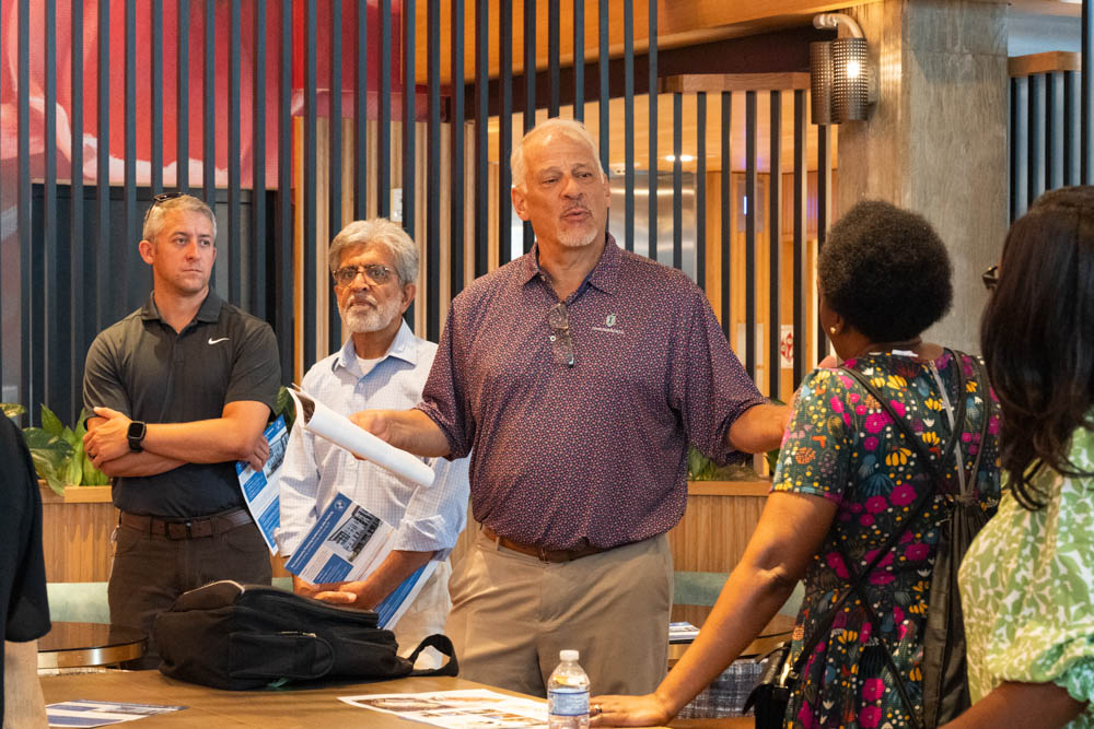 Five people stand in a modern indoor setting, engaged in conversation. One man gestures as he speaks, while others listen. Papers and a water bottle are on the table.