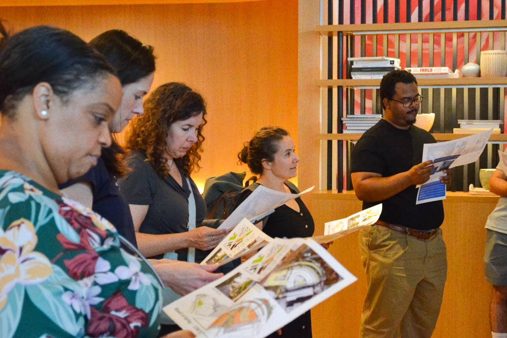 A group of people stands indoors, reading printed documents or maps, with shelves and books visible in the background.