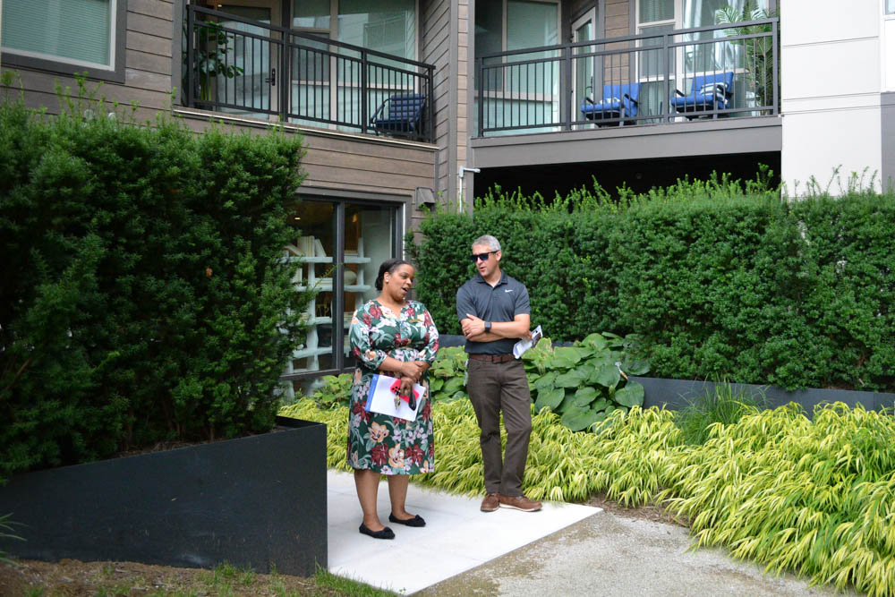 Two people stand on a walkway outside a modern apartment building, engaged in conversation, with greenery and bushes around them.