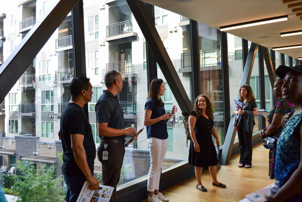A group of people stands and talks in a modern glass hallway with large windows overlooking an apartment building.