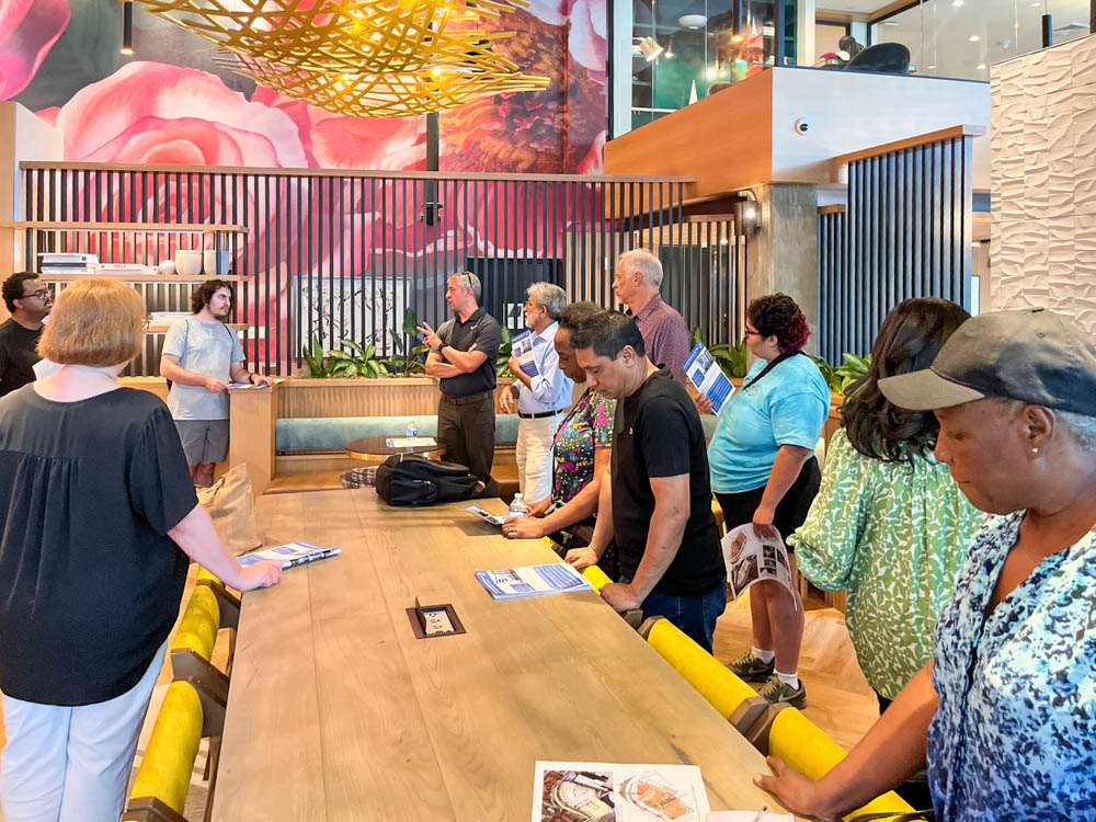 A group of people stands around a long wooden table in a modern, brightly decorated room, listening to a woman speaking. Some hold papers or pamphlets.
