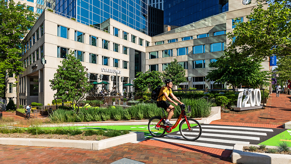A person wearing a backpack rides a red bicycle across a striped crosswalk in front of a modern office building with trees and outdoor seating.