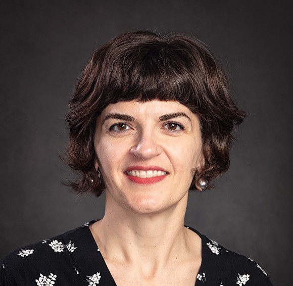 Woman with short, dark wavy hair and bangs, wearing pearl earrings and a dark patterned top, smiling in front of a plain, dark background.