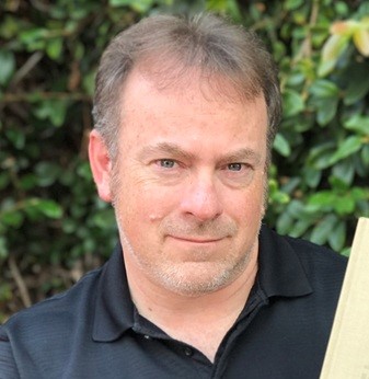 A man with short brown hair and a trimmed beard wearing a black polo shirt stands outdoors in front of green foliage.