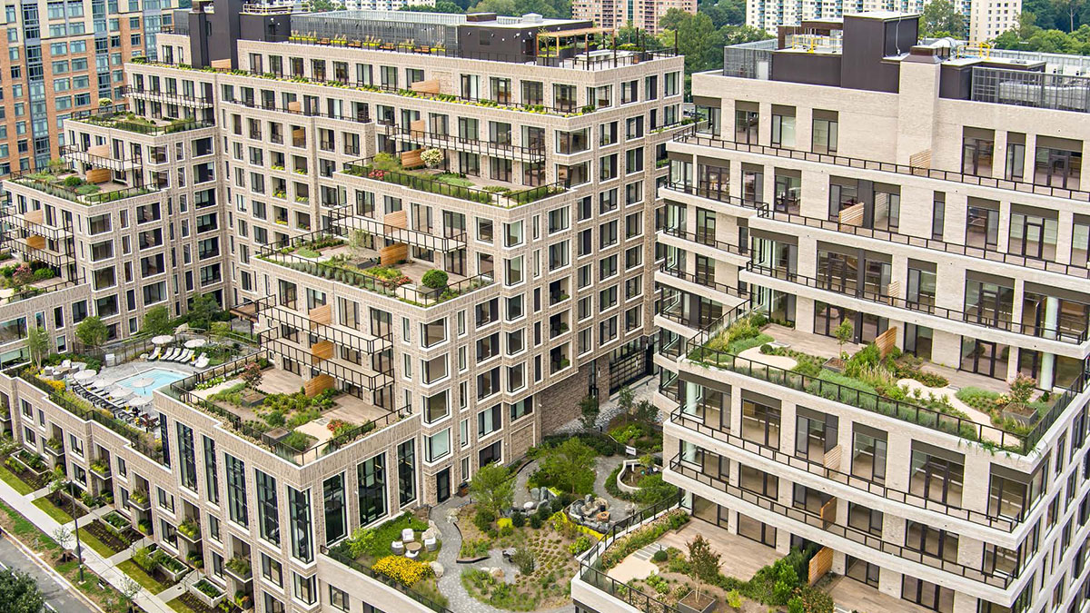 A modern multi-story apartment complex with large balconies, rooftop gardens, and outdoor terraces, surrounded by other buildings and greenery.
