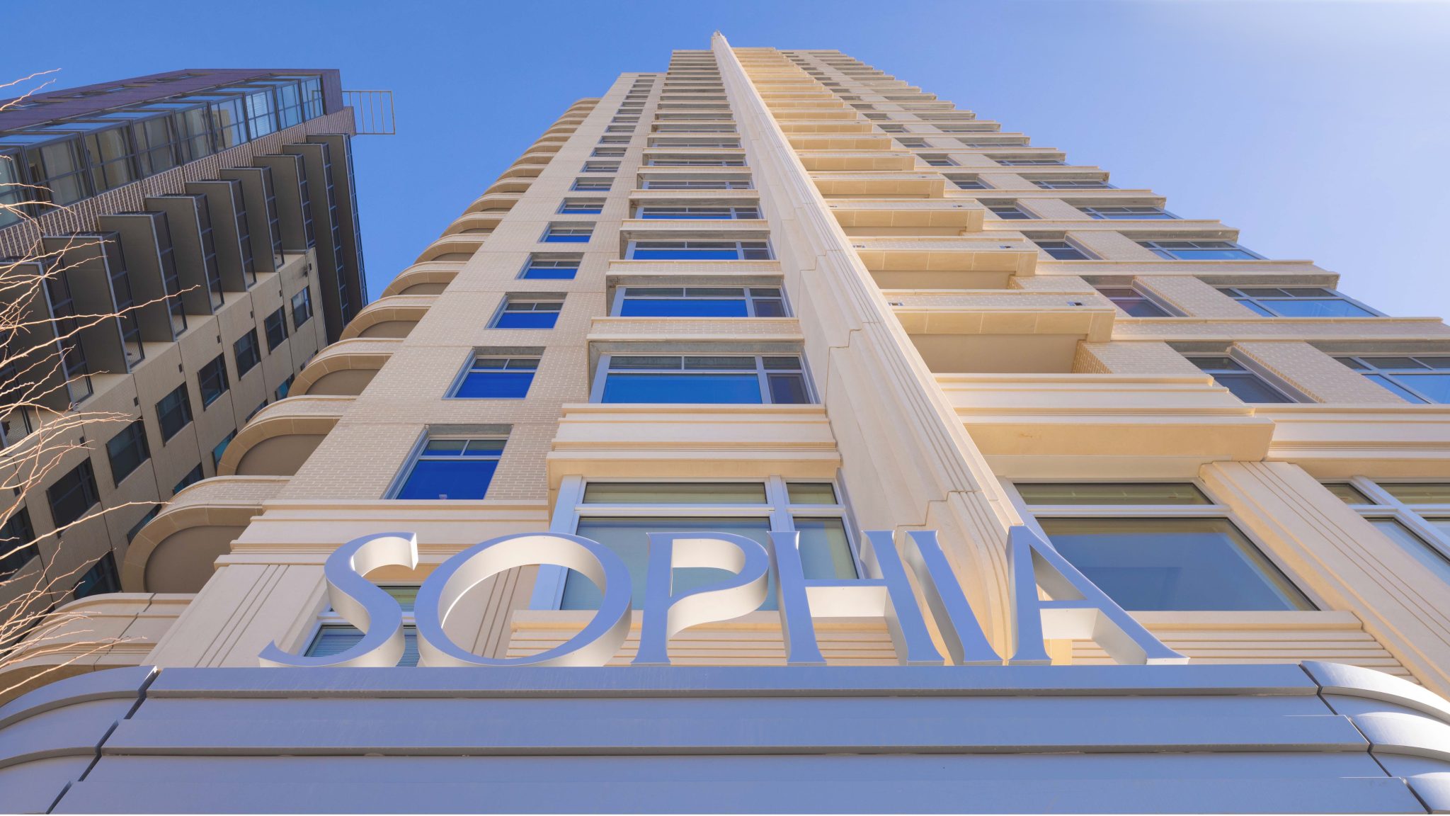 A tall beige apartment building viewed from below, with large windows and a sign reading "SOPHIA" at the entrance, set against a clear blue sky.