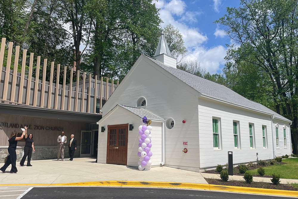 A small white church with green-trimmed windows and a steeple, decorated with purple and white balloons, stands beside a modern building. Several people are gathered outside.