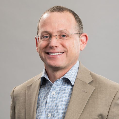 A man with short hair and glasses, wearing a beige suit jacket over a blue checked shirt, smiles in front of a plain gray background.