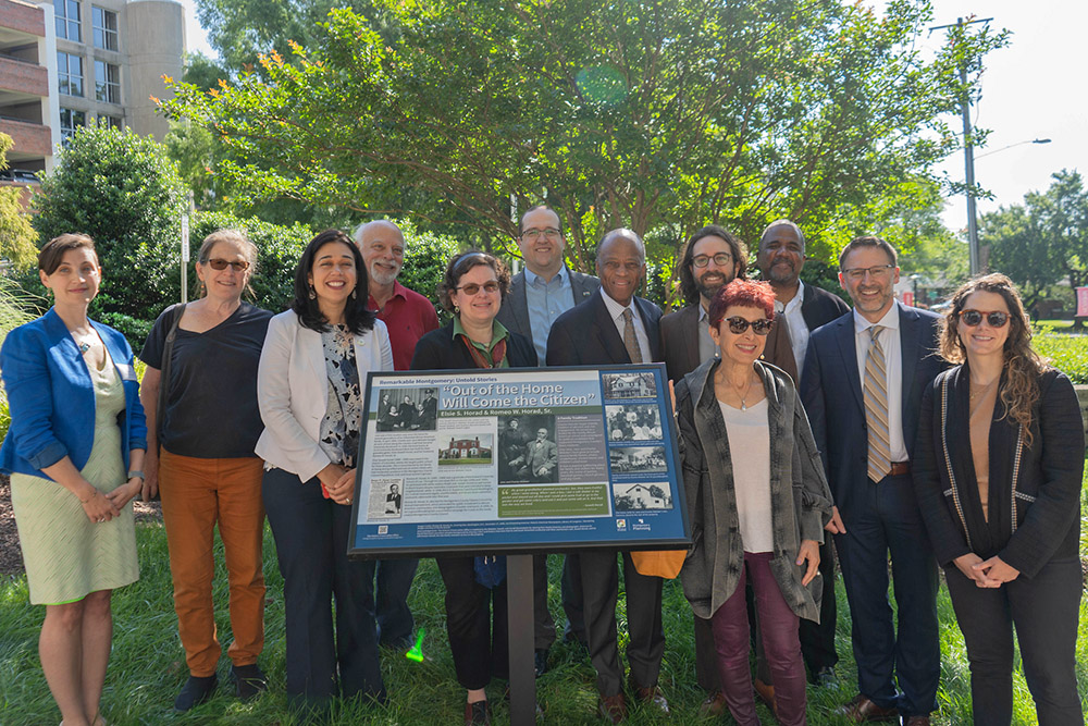 A group of thirteen people stands outdoors behind an informational sign, posing for a photo on a sunny day with trees and buildings in the background.