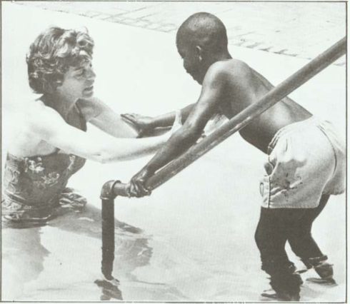 A woman in a swimsuit helps a young boy, who is standing on pool steps and holding a handrail, enter a swimming pool.