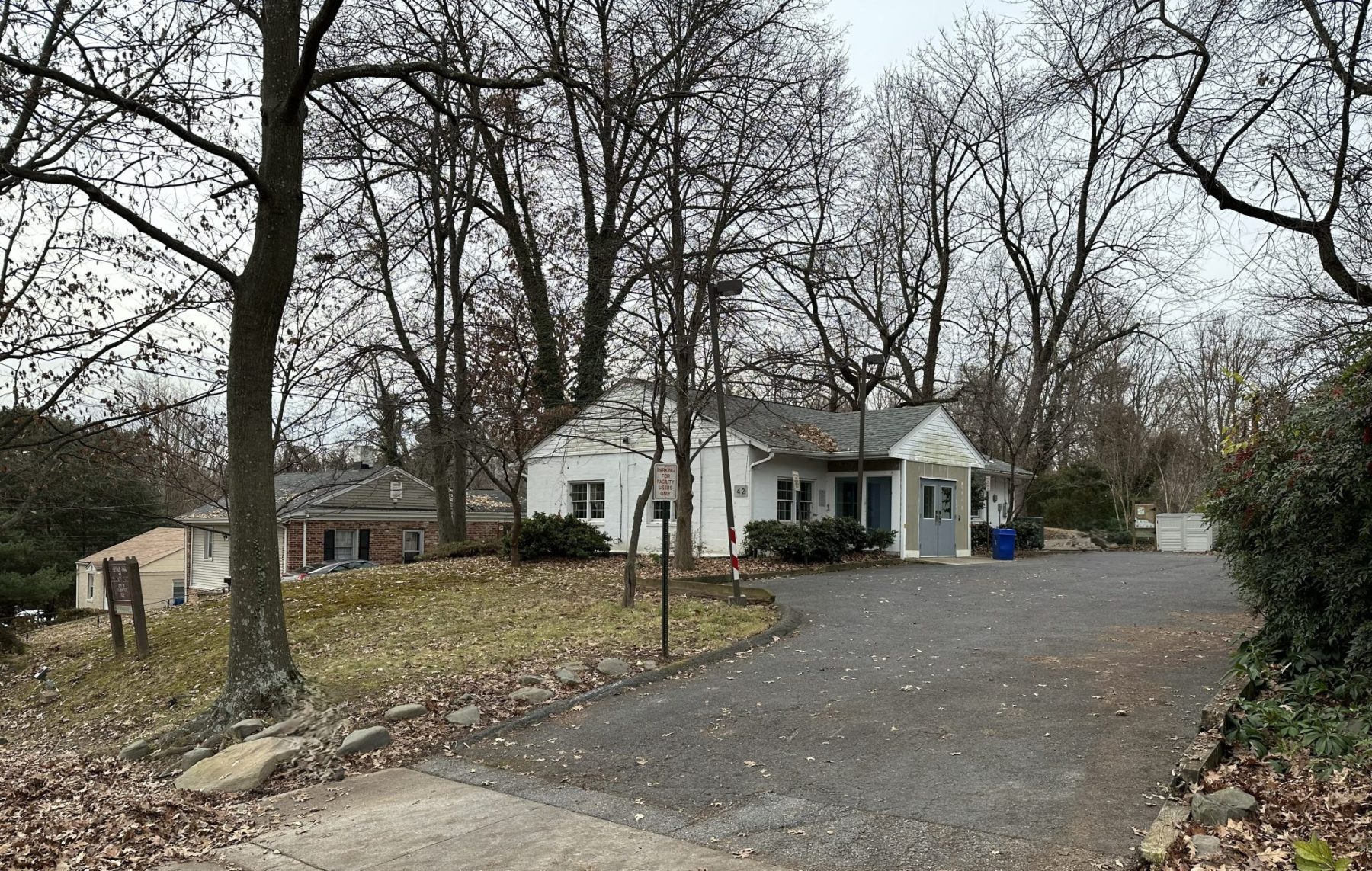 A small white house with a blue front door sits on a sloped lot with bare trees and a paved driveway on an overcast day.