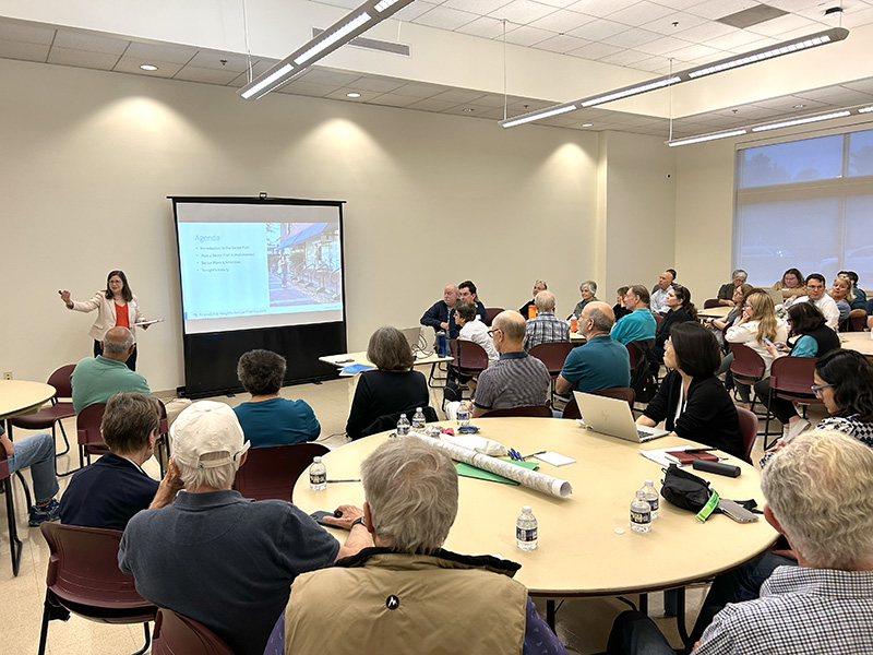 A woman presents slides to a group of people seated at round tables in a well-lit meeting room.