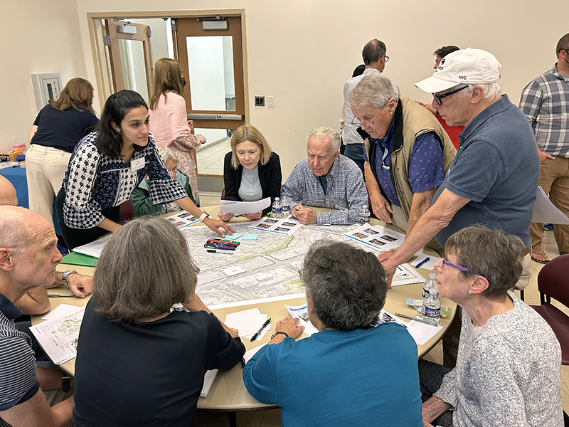 A group of adults gathers around a table covered with maps and papers, actively discussing and pointing at documents in a meeting room.