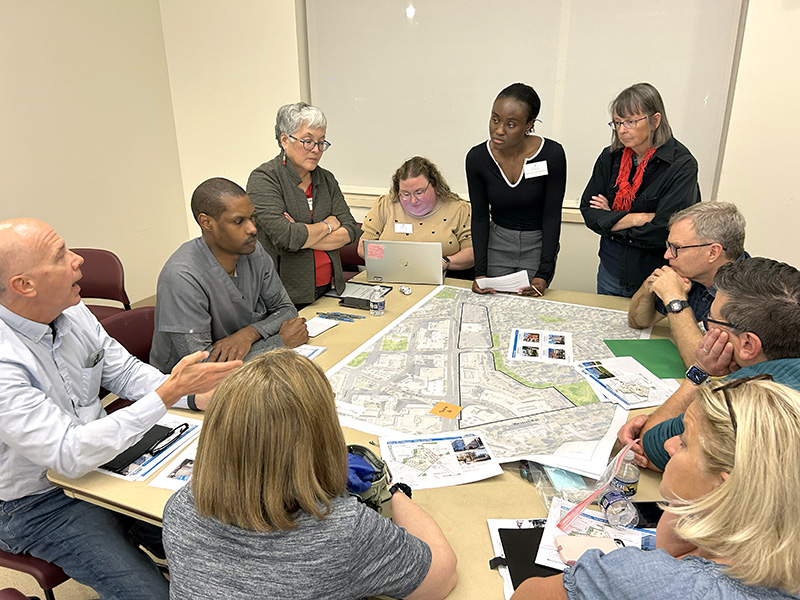 A group of people sit and stand around a table, discussing a large map and planning documents during a meeting.