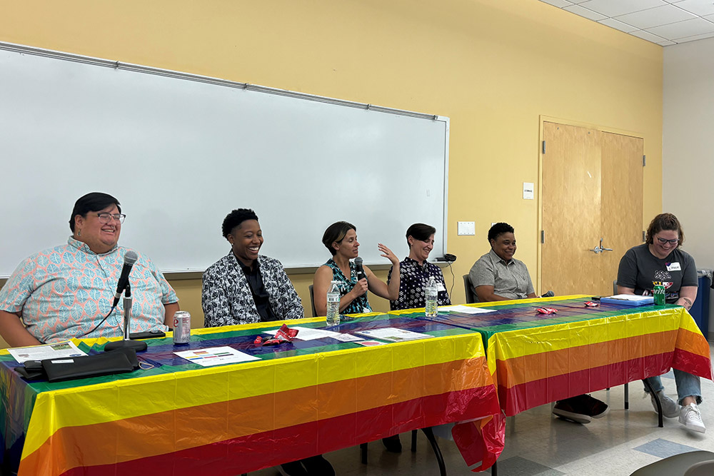 Six people sit at a table covered with a rainbow flag, participating in a panel discussion in a classroom setting.