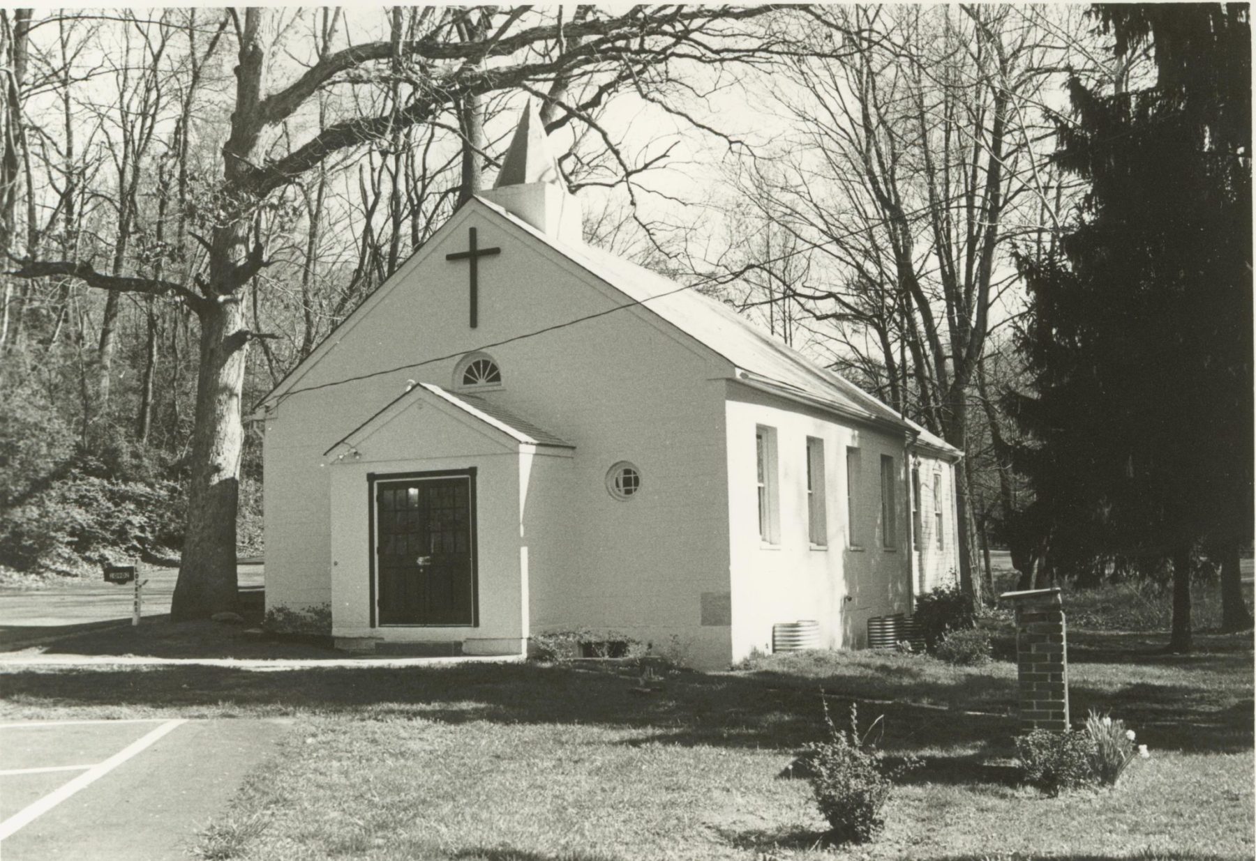 Black-and-white photo of a small, white church with a cross above the entrance, surrounded by leafless trees and a grassy area.